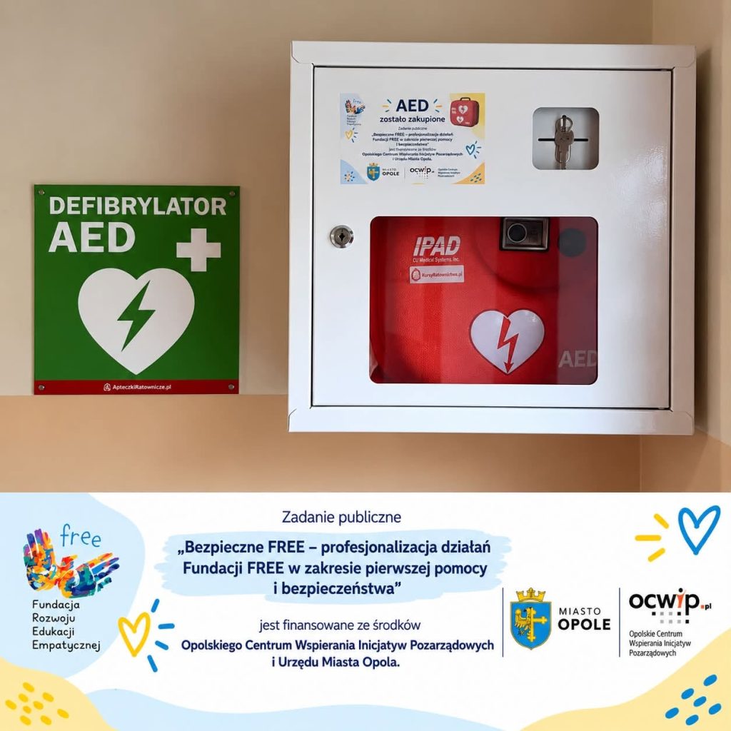 Defibrylator AED w białej szafce na ścianie. Obok zielone oznaczenie AED. Zakup urządzenia został sfinansowany przez Opolskie Centrum Wspierania Inicjatyw Pozarządowych i Urząd Miasta Opola.