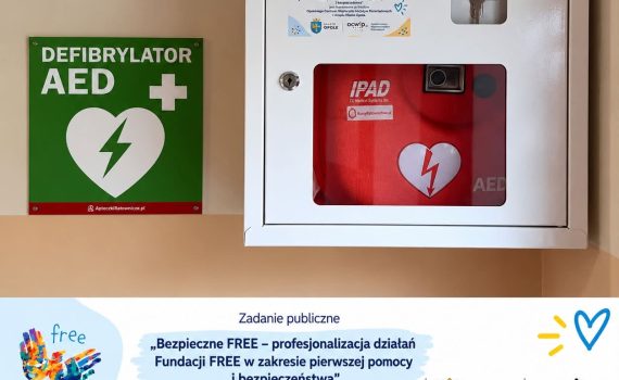Defibrylator AED w białej szafce na ścianie. Obok zielone oznaczenie AED. Zakup urządzenia został sfinansowany przez Opolskie Centrum Wspierania Inicjatyw Pozarządowych i Urząd Miasta Opola.